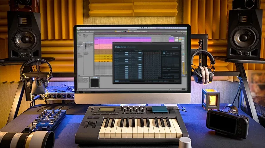 Escritorio de estudio con espuma acústica amarilla, dos monitores de estudio sobre pedestales, audífonos, teclado controlador MIDI y una pantalla con una sesión de DAW y la ventana del Dolby Atmos Composer abierta