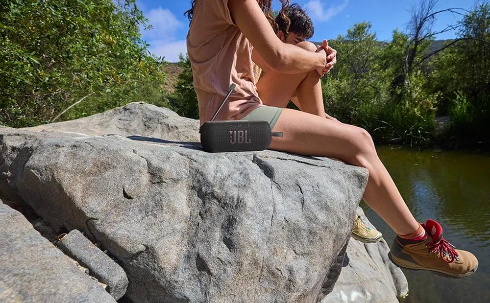 JBL Tuner 3 sobre una roca junto al río, con una persona sentada al lado